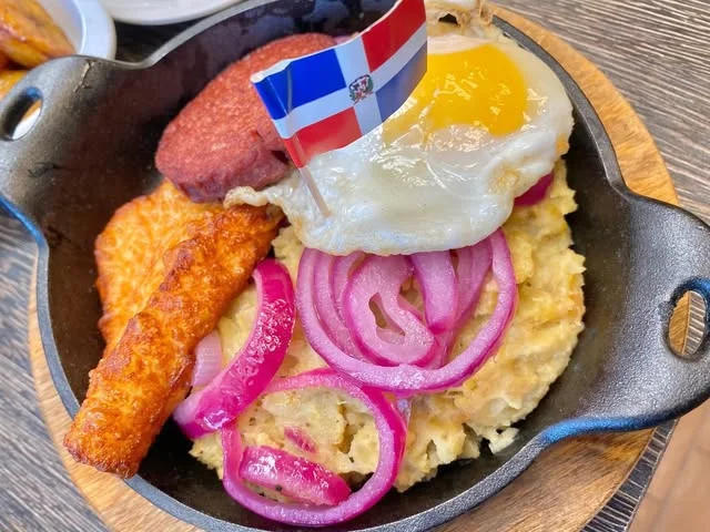 Mangú in a cast-iron skillet with a fried egg, salami, fried cheese and pickled red onion under a Dominican flag pick